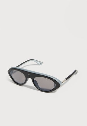 Gafas de sol estilo ojo de gato en negro y gris con lentes tintados oscuros, colocadas sobre una superficie lisa de color gris claro.