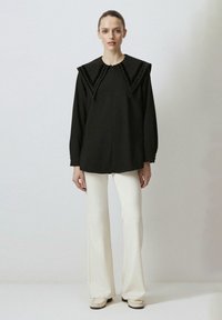 Camisetta nera a maniche lunghe con colletto strutturato, caratterizzata da dettagli verticali, abbinata a pantaloni bianchi a campana e scarpe beige senza lacci.
