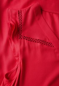 Czerwony top z tkaniny z falbaniastymi krawędziami i okrągłymi wycięciami. Gładka faktura, dekolt w kształcie litery V oraz przeszycia wzdłuż szwów.