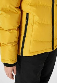 Žltá puffer bunda so zipsovou vreckom na boku, s hladkým povrchom a kontrastným čiernym zipsom a manžetami.