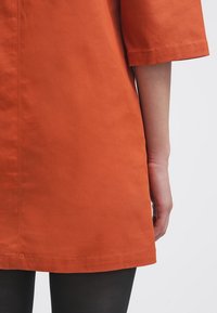 Haut en coton orange à manches trois quarts. Présente une coupe droite, des coutures visibles et une texture douce. Porté avec des leggings noirs.