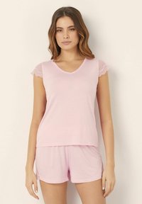Set de pijama roz cu mâneci scurte, prezentând un top cu decolteu în V și accente din dantelă pe umeri, împreună cu pantaloni scurți asortați, cu o croială lejeră. Textură moale a materialului.