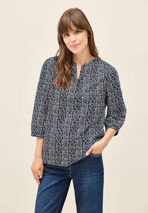 Blusa blu navy con un motivo geometrico bianco, caratterizzata da un colletto alto e maniche a tre quarti. Abbinata a jeans in denim blu.