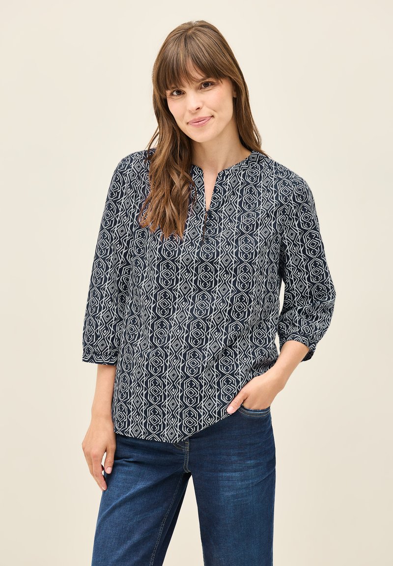 Blusa azul marino con un patrón geométrico blanco, con cuello alto y mangas de tres cuartos. Combinada con jeans de mezclilla azul.
