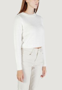 Donna che indossa un maglione bianco soffice a maniche lunghe e pantaloni beige a vita alta, mostrata dalla metà della spalla ai fianchi contro uno sfondo semplice.