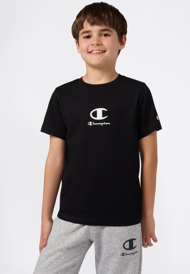 T-shirt nera in cotone con il logo "Champion" bianco al centro. Indossato con pantaloni della tuta grigi chiari che mostrano un logo abbinato.