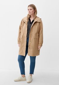 Hellbeige, knielanger Regenmantel mit Kapuze und Kordelzug, Druckkn�öpfen und zwei Fronttaschen, kombiniert mit blauen Jeans und weißen Turnschuhen.