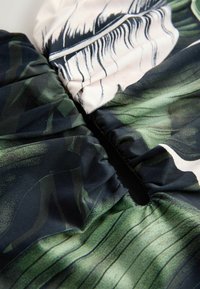 Tamsiai žalia ir juoda medžiaga su lapų raštu; turi sugriežtintą tekstūrą ir dalinai atvirą siūlę kaip dizaino užsegimą.