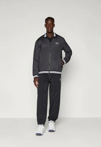 Jachetă gri cu fermoar, cu guler și manșete cu dungi albe, purtată împreună cu pantaloni asortați, ambele având un logo. Acesorii cu adidași albi.