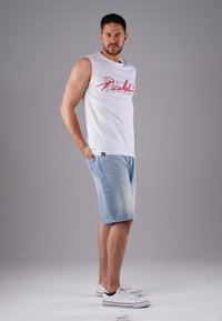 White sleeveless shirt with red text, denim shorts, and white sneakers with red accents, showcasing a casual summer outfit.