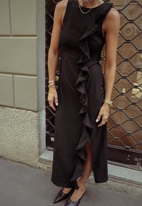 Musta olkaimeton mekko, jossa on röyhelökoristus sivulla ja etuaukko. Sujuva kankaan rakenne, yhdistettynä kärkitossuihin.