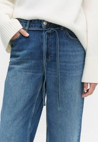 Ljusblå denimjeans med dragsko i midjan, med avslappnad passform och subtila sömdetaljer. Kombineras med en krämfärgad tröja.