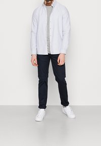 Ljusblå skjorta med knappar över en grå t-shirt, ihop med marinblå byxor och vita sneakers. Skjortan har en klassisk krage och långa ärmar.