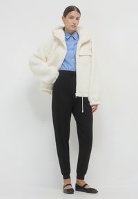 Weiße Fleecejacke mit Kapuze, mit einer Vordertasche, getragen über einem blau gestreiften Hemd und schwarzen Hosen. Schwarze flache Schuhe runden das Outfit ab.