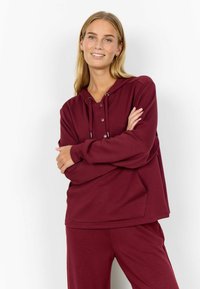Femme aux longs cheveux blonds portant un sweat à capuche bordeaux et un pantalon assorti, souriant avec les bras croisés sur un fond blanc.