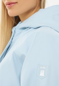 Lichtblauwe waterdichte jas met capuchon, met een gladde textuur en een witte logopatch op de linkerschouder.