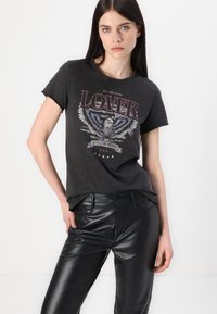 Donkergrijze crew-neck t-shirt met een afbeelding van een arend en de tekst "ALL AMERICAN LOVER" in het rood. Gedragen met glanzende zwarte leren broek.