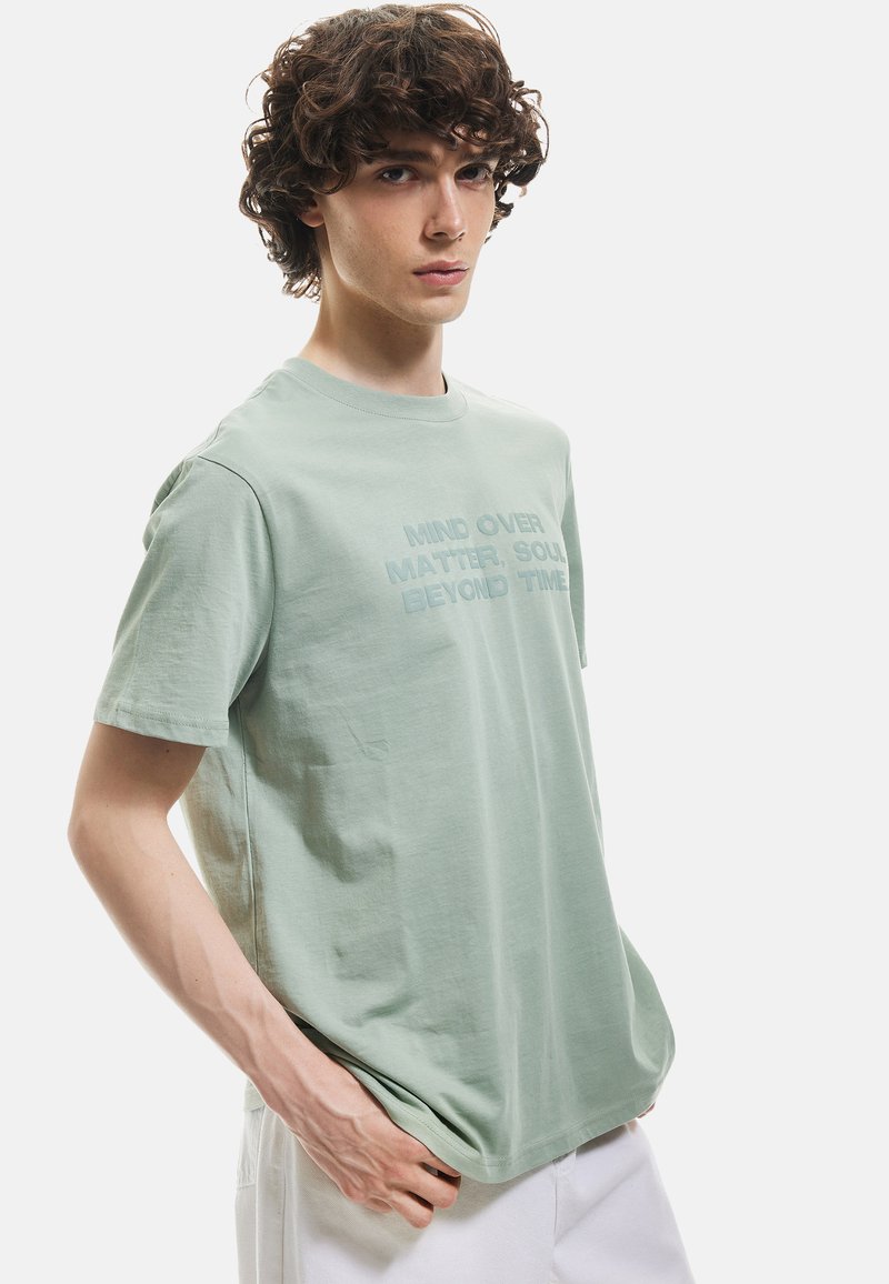 Camiseta de algodón verde claro con mangas cortas y cuello redondo. Presenta un estampado gráfico texturizado que dice "LA MENTE SOBRE LA MATERIA, EL ALMA MÁS ALLÁ DEL TIEMPO."