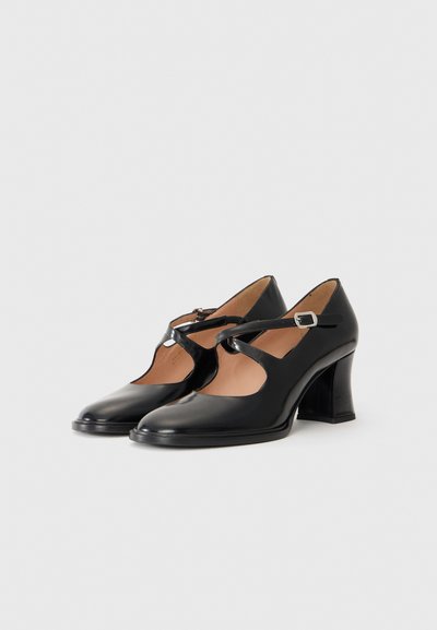Des chaussures à talons hauts en cuir noir avec un bout arrondi, des découpes sur les côtés et une fine bride à la cheville maintenue par une boucle en métal.