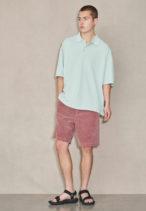 Fiatal férfi áll sima fal előtt, világoskék, túlméretezett pólót, porró rózsaszín rövidnadrágot és fekete, pántos szandált visel.