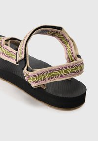 Flate sandal med svart gummi yttersåle, teksturert innersåle og vevd flerfarget tekstiloverdel med grønne, rosa og kremfargede mønstre.