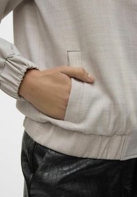 Ljust beige texturerad jacka med samlade manschetter, utrustad med en sidoficka. Matchas med svarta läderbyxor med krokodilpräge.