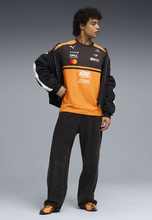 Junger Mann, der ein oranges und schwarzes Sportshirt mit Markenlogo trägt, eine schwarze Jacke über den Schultern trägt, lockere schwarze Hose und orangefarbene Sneakers trägt, steht und schaut nach rechts.