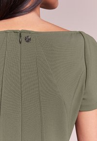 Oliwkowa bluzka o gładkiej fakturze, z krótkimi rękawami, szerokim kołnierzem i ukrytym zamkiem z tyłu z małym detalem w postaci logo.