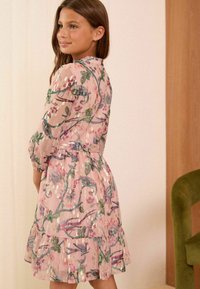 Abito con stampa floreale in tessuto semi-trasparente rosa con dettagli verdi. Presenta maniche lunghe, un orlo smerlato e una vita aderente.