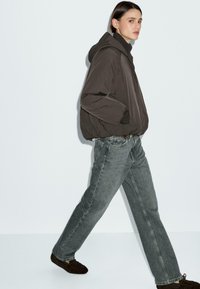 Persona che cammina indossando un giubbotto imbottito scuro, jeans grigi, dolcevita nera e mocassini scuri su uno sfondo chiaro uniforme.