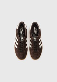 Brūnas zamšādas sporta kurpes ar trim baltām ādas svītrām, melnām šņorēm un gumijas zolē. Zīmola marķējums mēlē un sānos zelta krāsā.