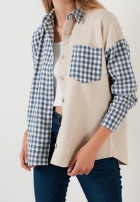 La camicia a maniche lunghe presenta un misto di lana beige da un lato e flanella di cotone blu a quadri dall'altro, con bottoni e una tasca.
