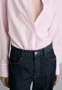 Camicia rosa chiaro con righe bianche e rosa più scuro, tasca frontale; abbinata a jeans denim scuri, cuciture arancioni e bottone in metallo.