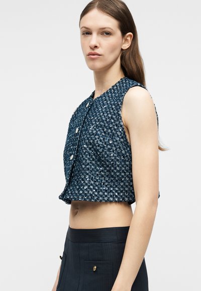 Νεαρή γυναίκα με μακριά καστανά μαλλιά, φορώντας αμάνικο cropped γιλέκο σε υφή σε ναυτικό μπλε χρώμα και ψηλόμεσα σκούρα παντελόνια, γυρισμένη στο πλάι.