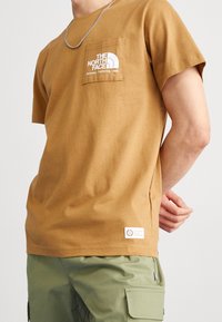 Brązowy t-shirt z krótkim rękawem wykonany z bawełny, z białym logo na kieszeni na piersi oraz metką na dole. Materiał o fakturze, luźny krój.