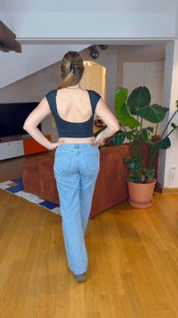 Top negro ajustado con escote en la espalda, combinado con jeans rectos de tiro medio en azul claro. Suelo de madera y plantas de interior visibles en el fondo.