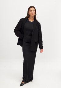 Mustan kohandatud bleiseri all on vormitud must kleit. Kleit on pikk ja voolujooneline, ning komplekti täiendavad terava ninaga mustad kontsad.