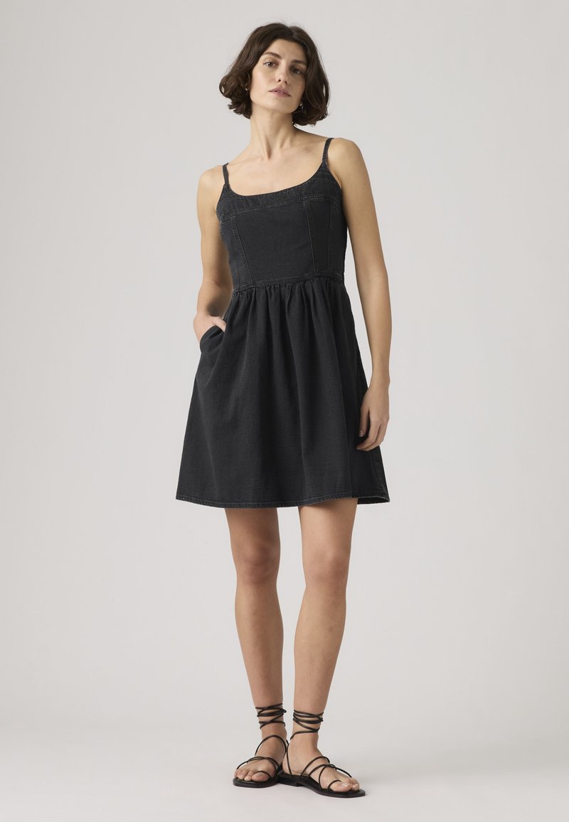 Vrouw staand met handen in de zakken, gekleed in een mouwloze zwarte jurk tot net boven de knieën en zwarte sandalen met bandjes en een platte zool tegen een effen achtergrond.