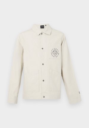 Giacca beige chiaro in cotone, con chiusura frontale a bottone, colletto e taschino sul petto con grafica del logo circolare. Due tasche frontali.