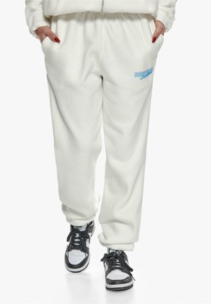 Weiße Fleece-Jogginghosen mit Seitentaschen, elastischem Bund und hellblauer Logobeschriftung. Kombiniert mit schwarz-weißen Sneakers.
