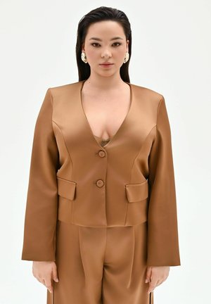 Femme portant une veste marron sur mesure avec un pantalon assorti et de grandes boucles d'oreilles en or, debout devant un fond blanc uni.