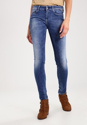 Vrouw die vervaagde blauwe skinny jeans en bruine enkellaarsjes draagt, staand tegen een effen achtergrond.