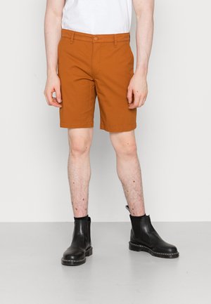 Mand iført rustfarvede shorts, hvid skjorte og sorte læderankelstøvler, der står mod en ensartet hvid baggrund.