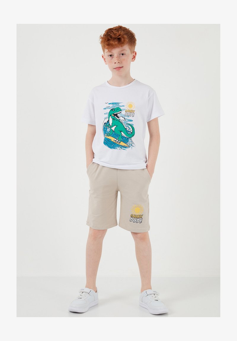 Garçon aux cheveux roux portant des baskets blanches, un short beige et un t-shirt blanc avec un dinosaure vert en train de surfer et le texte « Gnarly Dude ».
