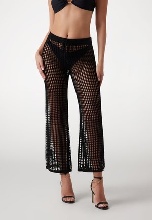 Schwarze Häkelhose mit weiter Beinform, die ein Gittermuster aufweist, sowie einem elastischen Bund und einer Kordelzugdetail. Getragen mit hochhackigen Sandalen.