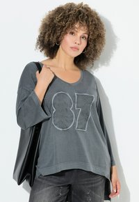Graues Langarm-T-Shirt mit V-Ausschnitt, versehen mit einer bestickten Nummer "87" und floralen Mustern. Kombiniert mit dunklen Jeans und einer schwarzen Tasche.