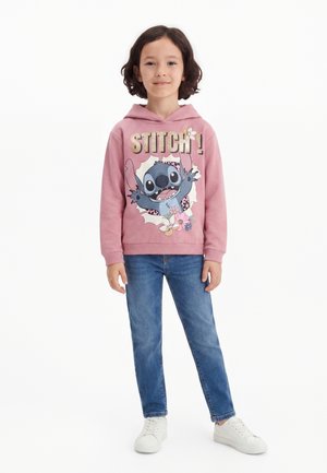 Barn iført en lyserød hættetrøje med en Stitch tegneserie og blå jeans, står med hænderne afslappet, smilende til kameraet mod en ensfarvet baggrund.
