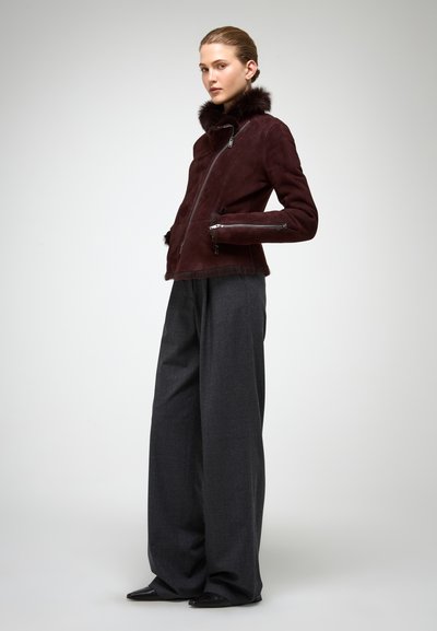 Mujer con chaqueta de ante burdeos con cuello y puños de piel, pantalones anchos oscuros y zapatos negros puntiagudos, posando de perfil contra un fondo liso.