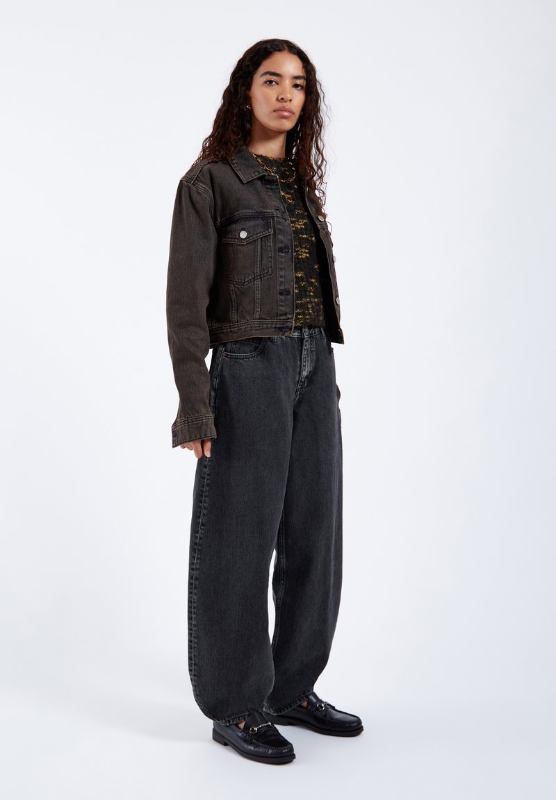 Junge Frau mit lockigem Haar trägt eine dunkle gekürzte Jeansjacke, ein gemustertes Top, weite dunkle Jeans und schwarze Loafers, stehend vor weißem Hintergrund.