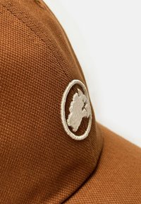 Gorra marrón hecha de tela texturizada, con un logo circular bordado en blanco que representa una silueta y un visera curva.